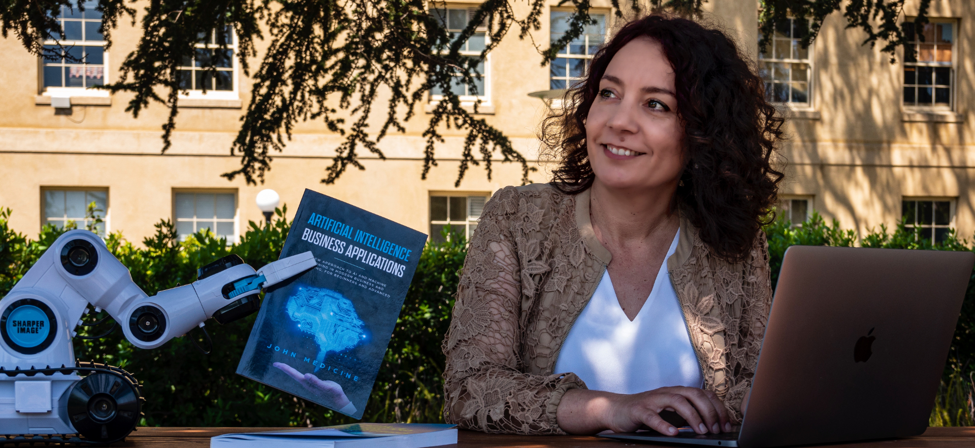 Carme Moreno seated outdoors with a laptop, next to a robot holding a book about artificial intelligence in business